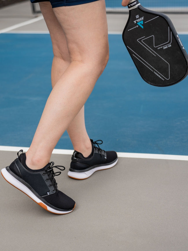 A woman holding a pickleball racquet on a tennis court wearing ATOM Women's Athletic Sneaker in color JetBlack-White-Gum