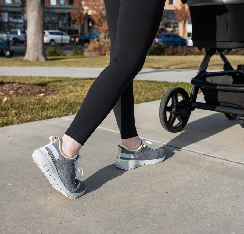 Woman strolling with her child in a baby carriage, showcasing her Dove Gray-Pale Lime FLEX Via women's sneakers by KURU Footwear, emphasizing the flexibility of the sole
