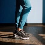 Person wearing black sneakers and teal pants standing on a textured surface with a blue wall in the background.