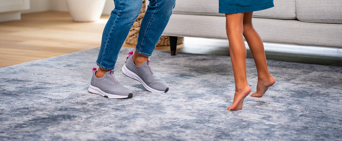 Two people at home jumping on a furry carpet. One of them is wearing comfortable indoor shoes by KURU Footwear, suitable for walking around the house