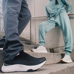 Two people on outdoor steps wearing KURU ATOM 2 Slip-On sneakers in Jet Black and Bright White.