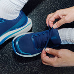 Close-up of hands tying a KURU DASH Vanta unisex sneaker in Indigo Blue on a gym floor.