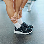 Person holding their calf in KURU DASH Vanta unisex sneakers in Jet Black-Charcoal at the gym.