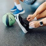 Person lacing a KURU DASH Vanta unisex sneaker in Jet Black beside a medicine ball at the gym.