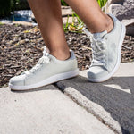 Woman stepping off a curb wearing KURU ROAM Women's Court Sneaker in Bone Gray-Cloud Gray.
