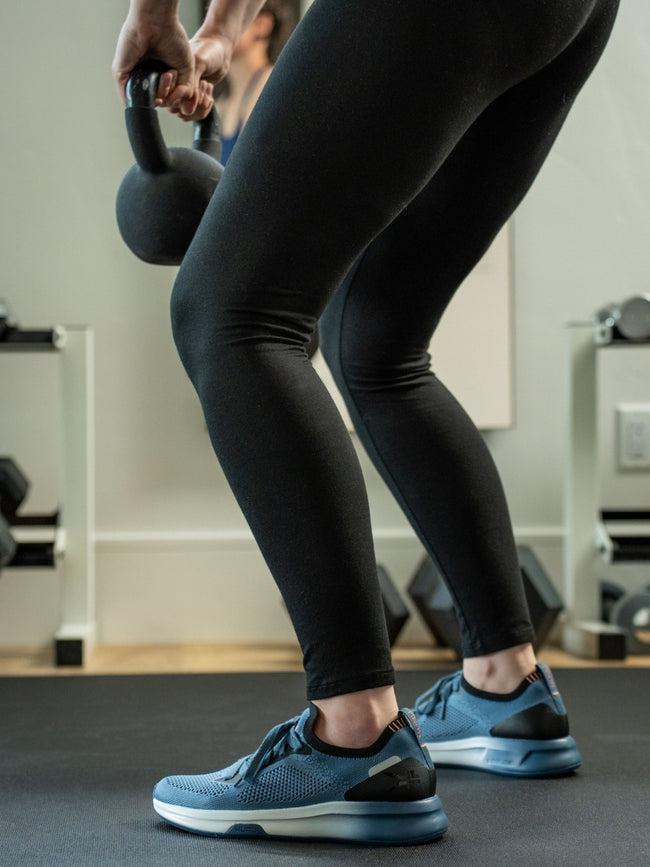 A woman lifting a kettlebell in a gym wearing KURU Footwear FLUX Women's Sneaker in MineralBlue-Black