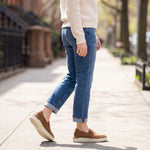 Person wearing blue jeans and brown shoes walking on a sidewalk.