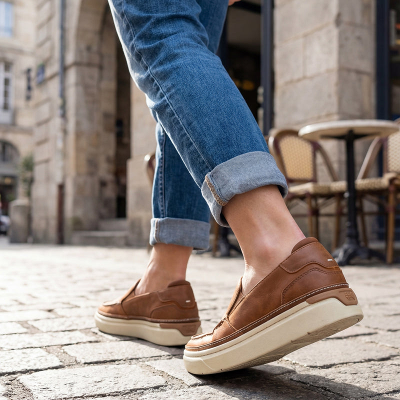 Brown slip-on shoes worn with rolled-up blue jeans on a street.