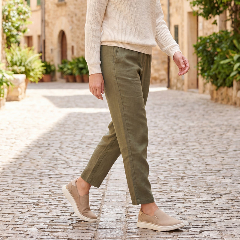 Person wearing a cream sweater, green pants, and beige shoes walking on a stone path.