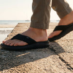 Person wearing black sandals on a stone surface with a blurred beach background