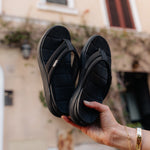 Black sandals held by a hand with a blurred outdoor background