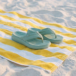 Pair of light blue sandals on a yellow and white striped towel on a sandy beach.