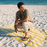 Man sitting on a yellow and blue striped towel on a sandy beach with ocean in the background