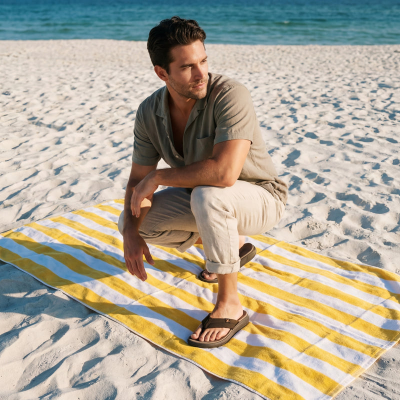 Man sitting on a yellow and blue striped towel on a sandy beach with ocean in the background