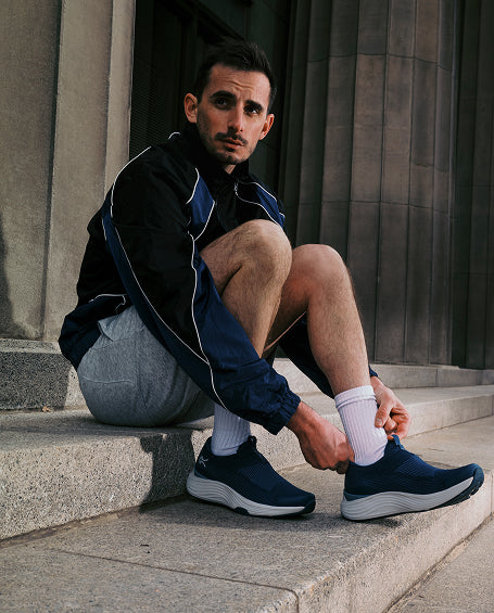 Man sitting on steps wearing a black jacket, gray pants, and blue sneakers.