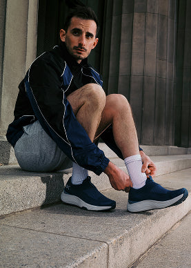 Man sitting on steps wearing a black jacket, gray pants, and navy running shoes.