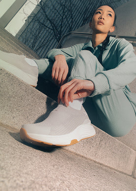 Person sitting on steps wearing a light green outfit and white sneakers with a brown sole.
